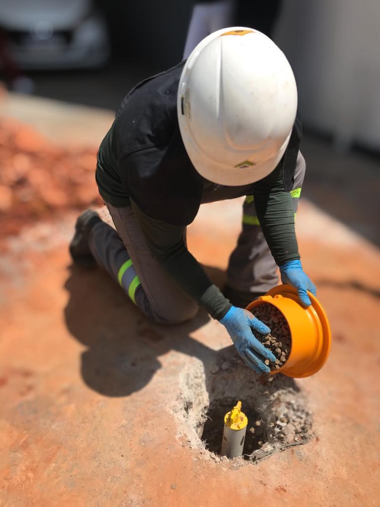 Técnico de joelhos insere material selante em furo de sondagem durante campanha de amostragem de solo da Ambsolution em campo
