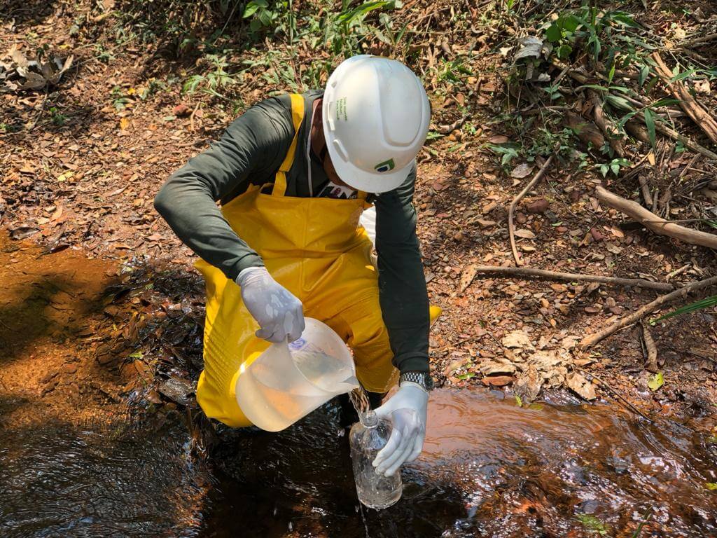 Profissional com EPI da Ambsolution Engenharia Ambiental coleta amostra de água em riacho para estudos técnicos de licenciamento ambiental
