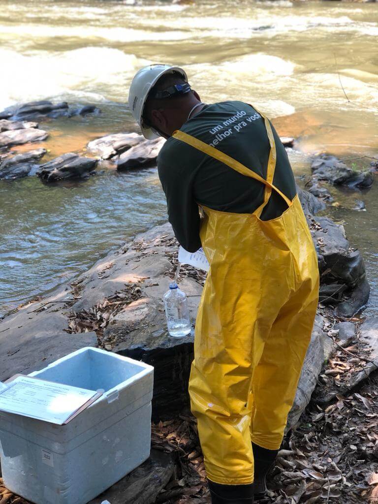 Técnico da Ambsolution com macacão amarelo coleta água de rio para monitorar projetos de reflorestamento e compensação ambiental